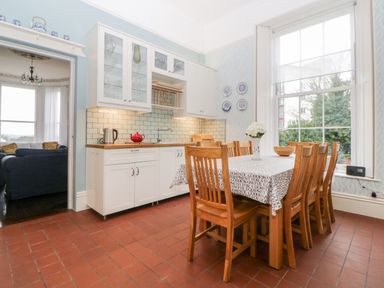 A kitchen with a wooden dining table and chairs near a large window and white cabinets with glass doors at Holcombe House in Torquay