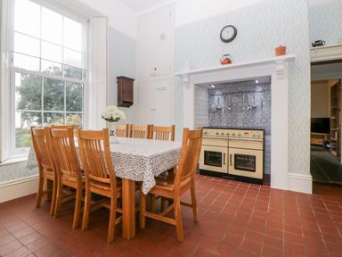 A dining room with a long wooden table and eight chairs a stove with patterned tiles behind it and a large window at Holcombe House in Torquay