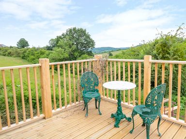 A wooden deck with two green metal chairs and a round table overlooking fields and trees at The Hideout in Llangrove