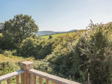 A wooden railing overlooking bushes, trees, green fields, and hills under a clear sky at The Hideout in Llangrove