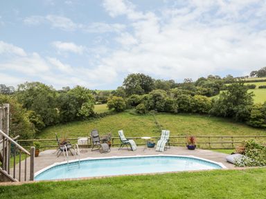 An outdoor swimming pool with lounge chairs and tables on a wooden deck overlooking green fields and trees at The Hideout in Llangrove