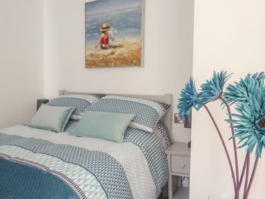 A bedroom with a bed and patterned bedding a bedside table and a painting of a child at the beach at Harbour View Apartment in Looe