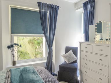 A bedroom with blue curtains a blue covered chair a white dresser with lighted mirror and a bed with blue bedding at Harbour View Apartment in Looe