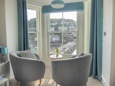 Two grey chairs with cushions and a small round table with a bottle and glasses by a window overlooking a bridge and houses at Harbour View Apartment in Looe