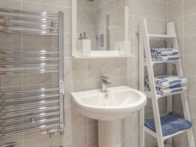 A bathroom with a white pedestal sink a chrome towel radiator a mirror above the sink and a white shelving unit with folded towels at Harbour View Apartment in Looe