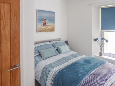 A bedroom with a double bed covered with blue and white bedding a wooden door a window with a blue blind and a framed painting on the wall at Harbour View Apartment in Looe