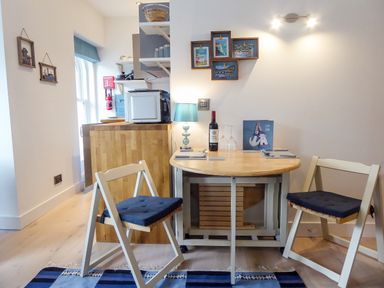 A small dining area with a round table, two chairs, a table lamp, wine bottle, and wall art at Harbour View Apartment in Looe