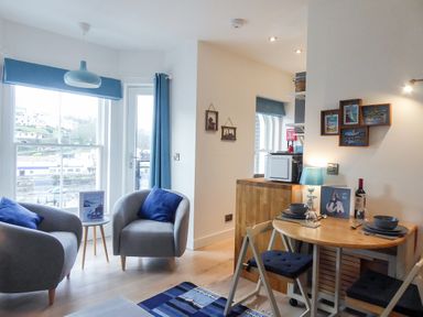 A sitting area with two gray armchairs and blue cushions near a window and door next to a small wooden dining table with two chairs and blue cushions at Harbour View Apartment in Looe