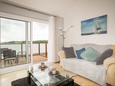 A living room with a sofa pillows a glass coffee table and sliding doors opening to a balcony overlooking water at Delfryd in Y Felinheli