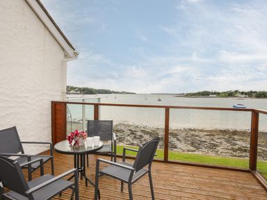 An outdoor deck with four chairs and a glass table overlooking a rocky shoreline and water at Delfryd in Y Felinheli