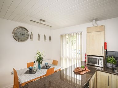 A kitchen with a dining table set with flowers and glasses a clock on the wall and a countertop with bread and vegetables at Delfryd in Y Felinheli