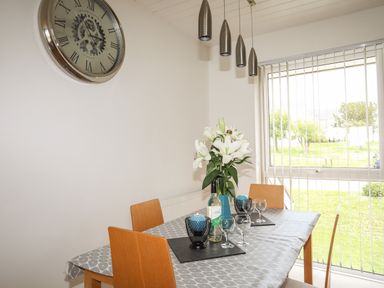 A dining area with a table covered by a tablecloth four wooden chairs a large wall clock a vase with flowers and a window with vertical blinds at Delfryd in Y Felinheli