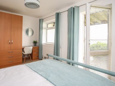 A bedroom with a bed, wooden wardrobe, desk with chair, round mirror, blue curtains, and a door with blinds at Delfryd in Y Felinheli