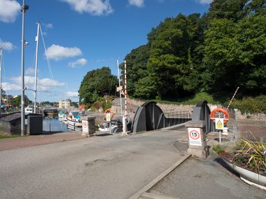 A metal swing bridge over water with sailboats docked beside it and trees in the background at Delfryd in Y Felinheli