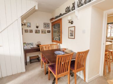 A dining area with a wooden table and four chairs next to a small desk under a staircase with framed pictures on the walls at Brackenbury Cottage in Beddgelert