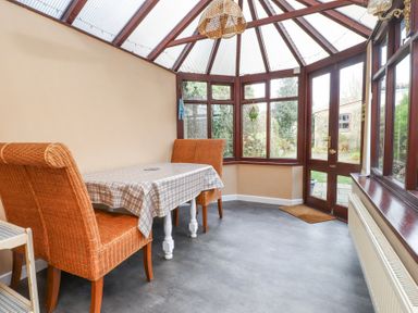 A dining area with wicker chairs and a table covered with a checkered tablecloth in a conservatory with glass windows and a door showing a garden at Brackenbury Cottage in Beddgelert