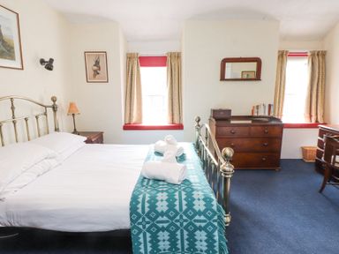 A bedroom with a metal bed frame towels on bed a wooden dresser mirror bookcase and two windows with curtains at Brackenbury Cottage in Beddgelert