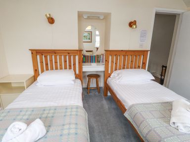 A bedroom with two single wooden beds with white linens and patterned blankets a wooden stool a built in desk with books and an oval mirror at Brackenbury Cottage in Beddgelert