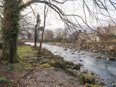 A river with rocks along the bank and trees on the left with stone houses on the right at Brackenbury Cottage in Beddgelert