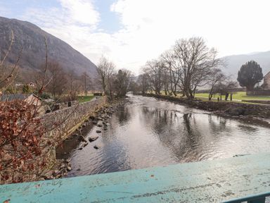 A river with stone walls and leafless trees along the banks with a mountain in the background at Brackenbury Cottage in Beddgelert