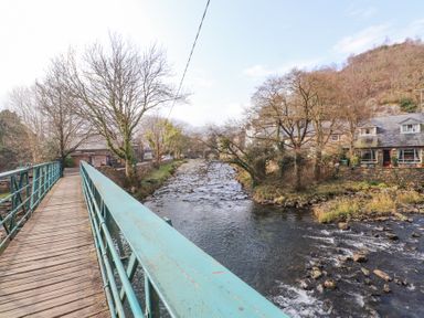 A wooden footbridge with green metal railings over a flowing river with houses and trees nearby at Brackenbury Cottage in Beddgelert