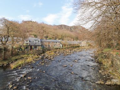 A river flowing alongside stone cottages with bare trees and hills in the background at Brackenbury Cottage in Beddgelert