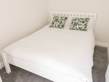A white bed with two green leaf patterned pillows in a room with white walls and gray carpet at Y Gogarth in Benllech