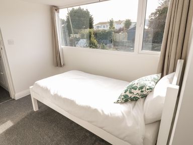 A bedroom with a single bed white bedding and a pillow with leaf pattern near a window with beige curtains at Y Gogarth in Benllech