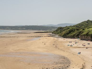 A wide sandy beach with scattered groups of people and cliffs with vegetation at Y Gogarth in Benllech