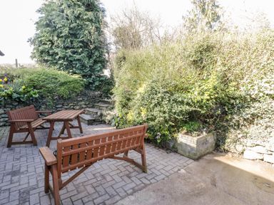 An outdoor patio area with a wooden bench and table surrounded by greenery and shrubs at Y Gogarth in Benllech