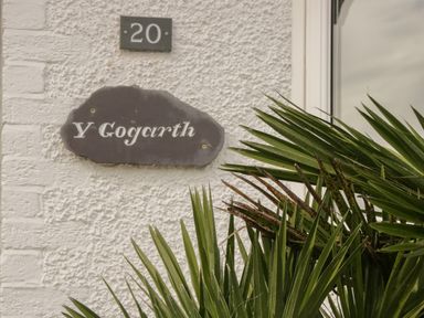 A house number 20 and a name plaque Y Gogarth on a textured wall with green palm leaves in front at Y Gogarth in Benllech