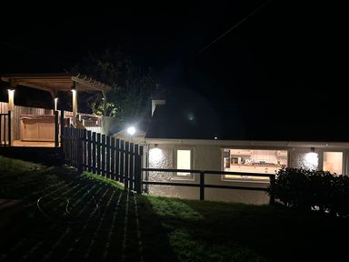A backyard at night with a wooden pergola hot tub and a house with illuminated kitchen windows at Y Gogarth in Benllech