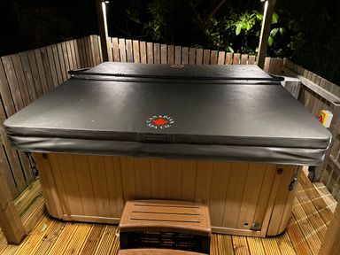 A covered hot tub on a wooden deck with small steps surrounded by a wooden fence at Y Gogarth in Benllech