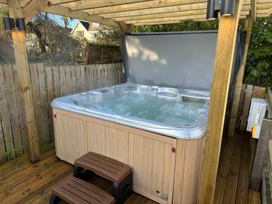 An outdoor hot tub with water bubbling under a wooden pergola with wooden steps in a fenced garden at Y Gogarth in Benllech