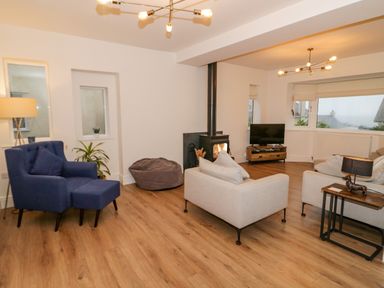A living room with a blue armchair, beige sofas, a wood stove, a television on a wooden stand, and wooden flooring at Y Gogarth in Benllech
