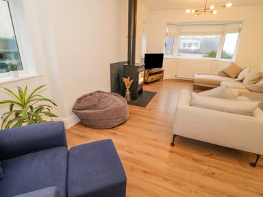 A living room with a blue armchair, a beige sectional sofa, a bean bag, a wood stove, a wooden tv stand with a television, plants, and large windows at Y Gogarth in Benllech