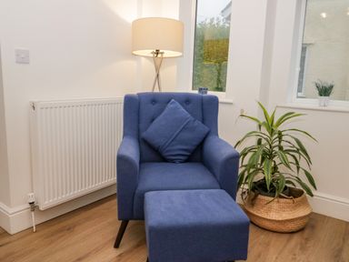 A blue armchair with a matching footstool and cushion next to a lamp radiator and a plant in a basket at Y Gogarth in Benllech