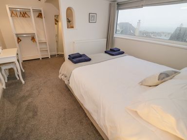 A bedroom with a bed, open wardrobe with hangers, a desk with stools, and a window showing rooftops at Y Gogarth in Benllech