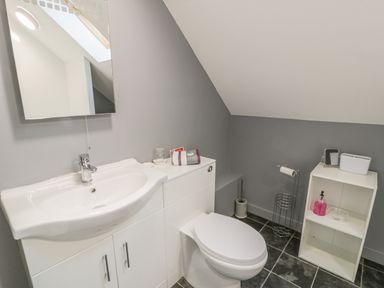 A bathroom with a white sink and cabinet a white toilet a small white shelving unit with a bottle of soap and toilet paper holder on a black tiled floor at Marchlyn in Deiniolen