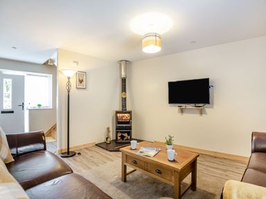 A living room with a wood stove a wall-mounted TV a wooden coffee table two leather sofas and a floor lamp at Marchlyn in Deiniolen