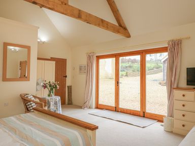 A bedroom with a bed a wicker chair a small table with flowers wooden framed glass doors and exposed wooden beams at The Apple Barn at Wyke Farm Lyme Regis