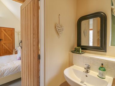 A bathroom sink with a mirror above and toiletries next to an open wooden door showing a bedroom at The Apple Barn at Wyke Farm in Lyme Regis