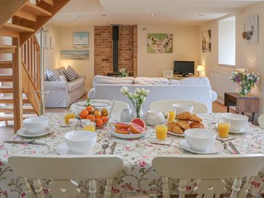 A dining table set with bowls glasses of orange juice croissants grapefruit and flowers with sofas a wood stove and paintings in the background at The Apple Barn at Wyke Farm Lyme Regis