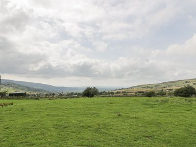 A grassy field with distant hills and scattered trees under a cloudy sky at Ffynnon Bach in Deiniolen