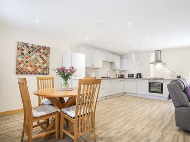 A kitchen area with white cabinets a dining table with four chairs and flowers on the table at Ffynnon Bach in Deiniolen
