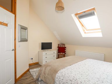 A bedroom with a bed a white dresser with a television a wooden chair with a red cushion a skylight window and a white door at Ffynnon Bach in Deiniolen
