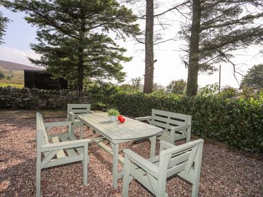 An outdoor seating area with a table and four chairs on gravel surrounded by trees and bushes at Ffynnon Bach in Deiniolen