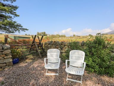 Two white chairs on gravel next to a stone wall with wooden steps leading over the wall at Ffynnon Bach in Deiniolen