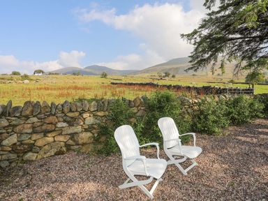 Two white chairs on gravel next to a stone wall with green fields and hills in the background at Ffynnon Bach in Deiniolen