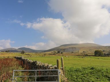 A field with a stone wall and a metal gate with hills and mountains in the background at Ffynnon Bach in Deiniolen